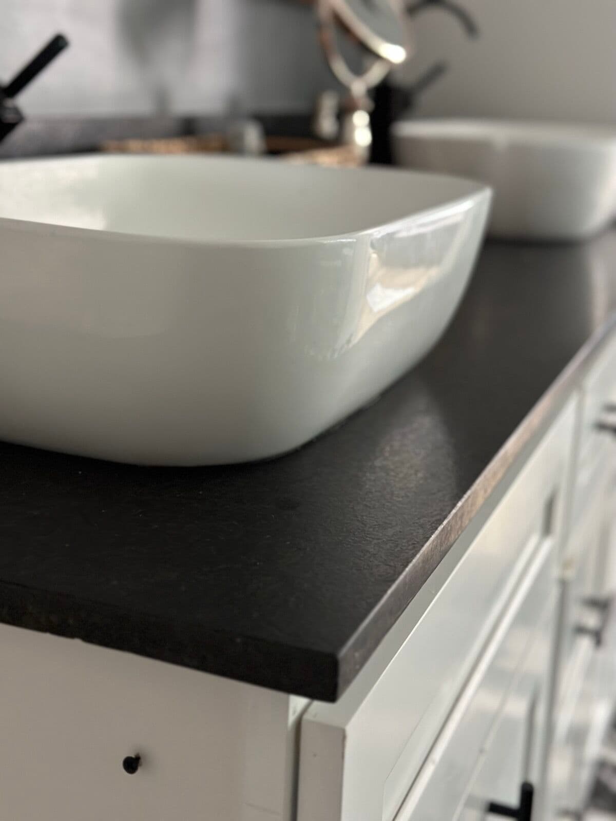 Close-up of a white rectangular vessel sink on a honed black countertop with white shaker drawers below