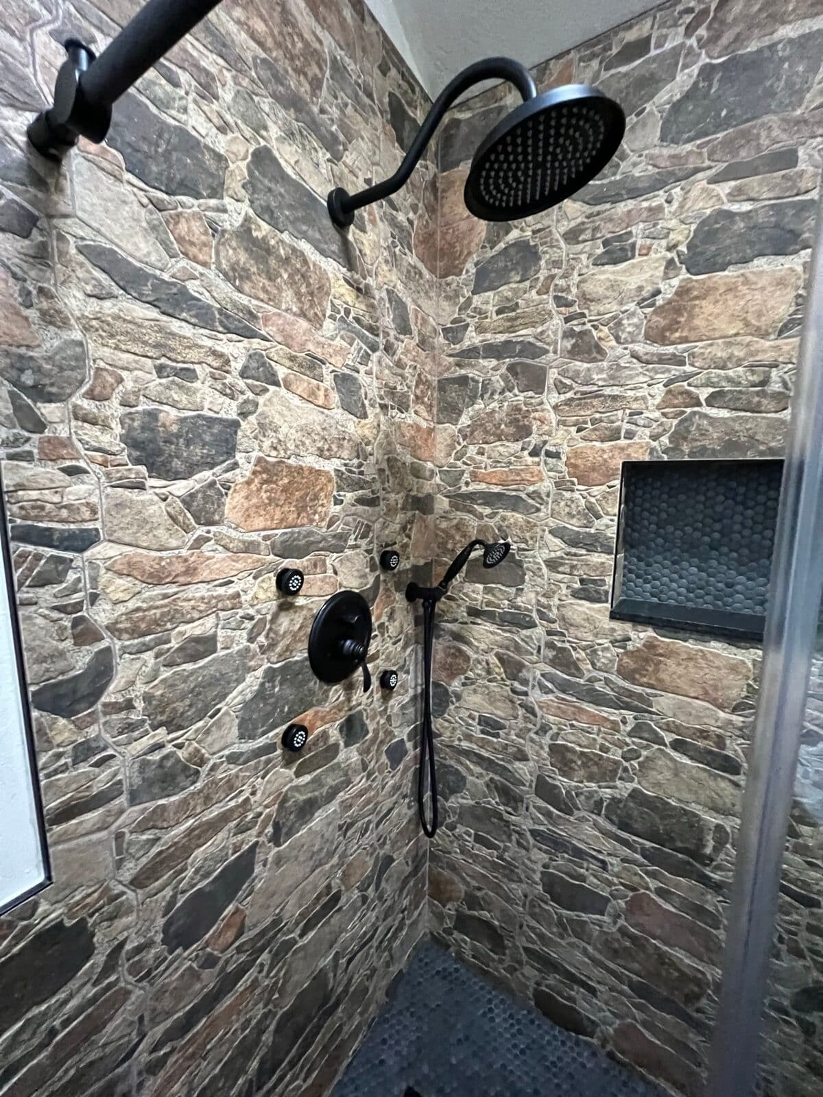 Matte-black rain shower head, hand shower, valve trim, and body sprays on stone-look tile walls