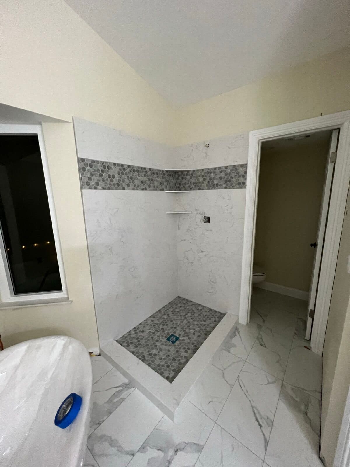 Curbed shower with marble-pattern wall panels, gray hex mosaic band and pan, next to a covered freestanding tub