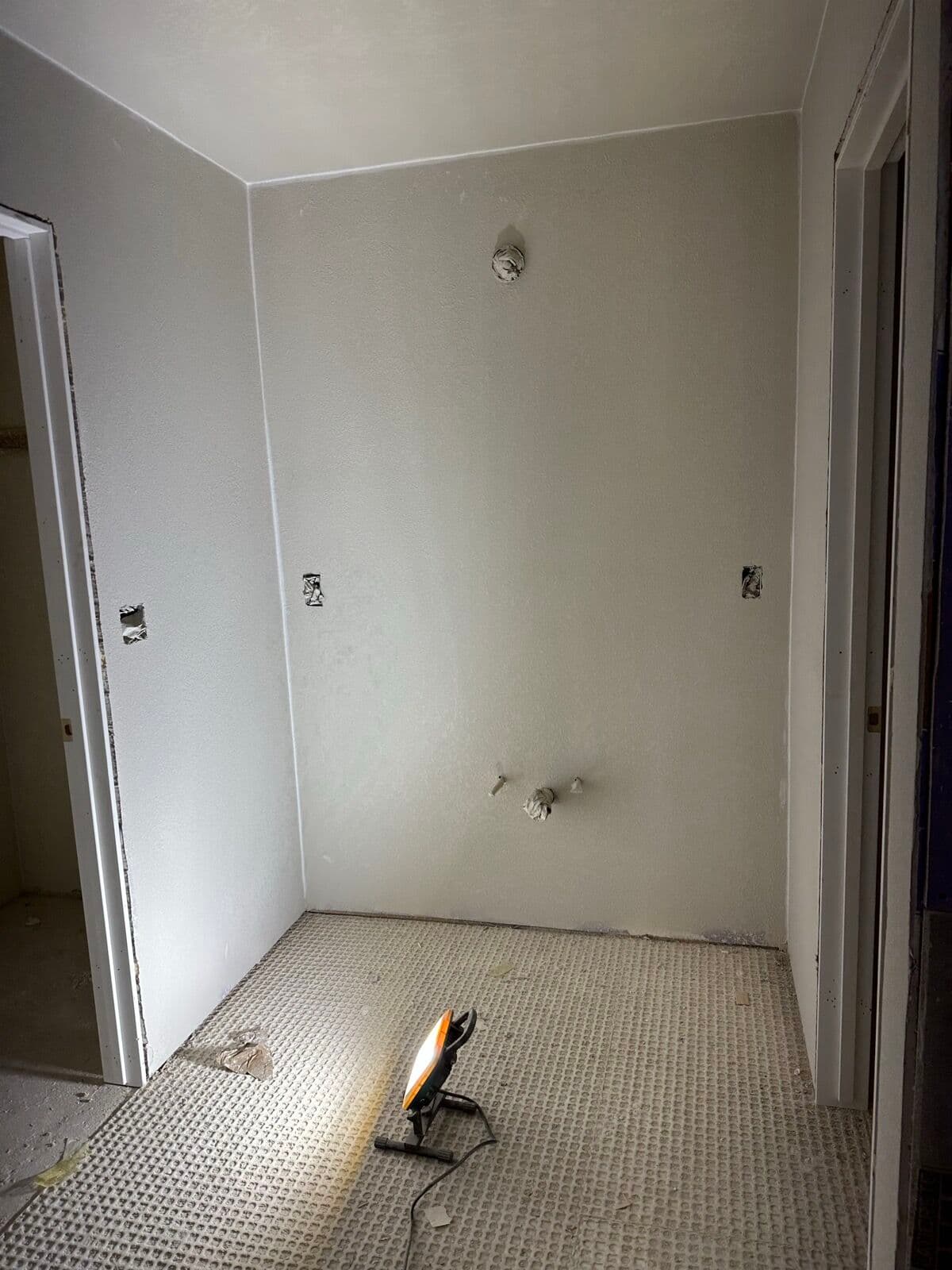 Bathroom with primed walls, an uncoupling membrane on the floor, a rough-in shower valve, and a work light