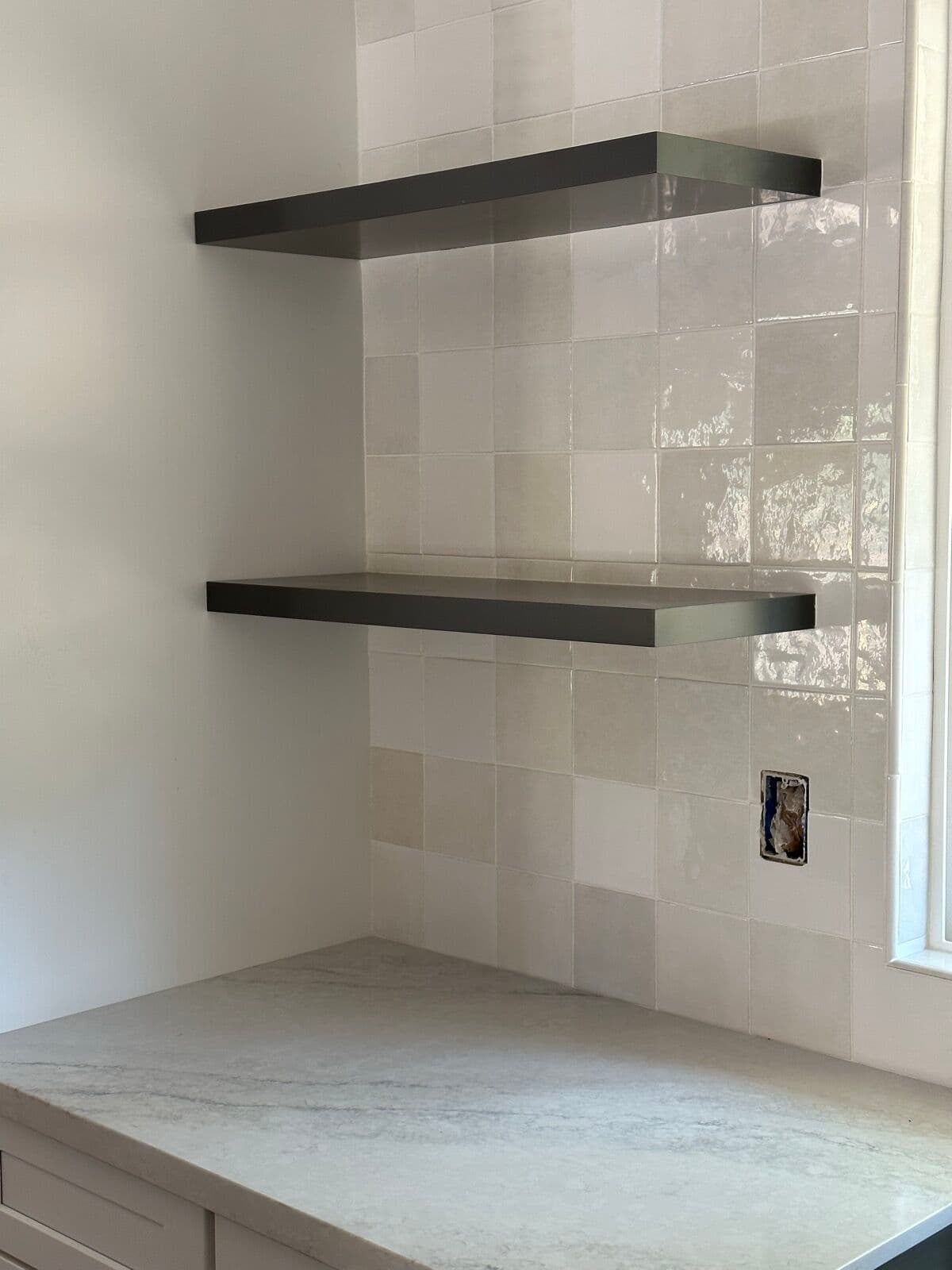 Two charcoal floating shelves mounted on a glossy square tile wall above a marble-look countertop
