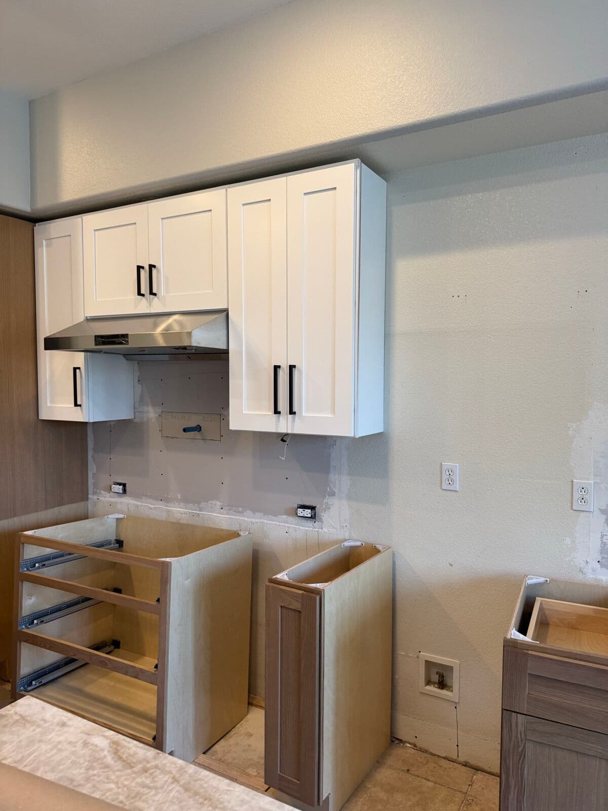 Kitchen mid-installation with white upper cabinets and hood mounted and unfinished oak base cabinet boxes with drawer glides