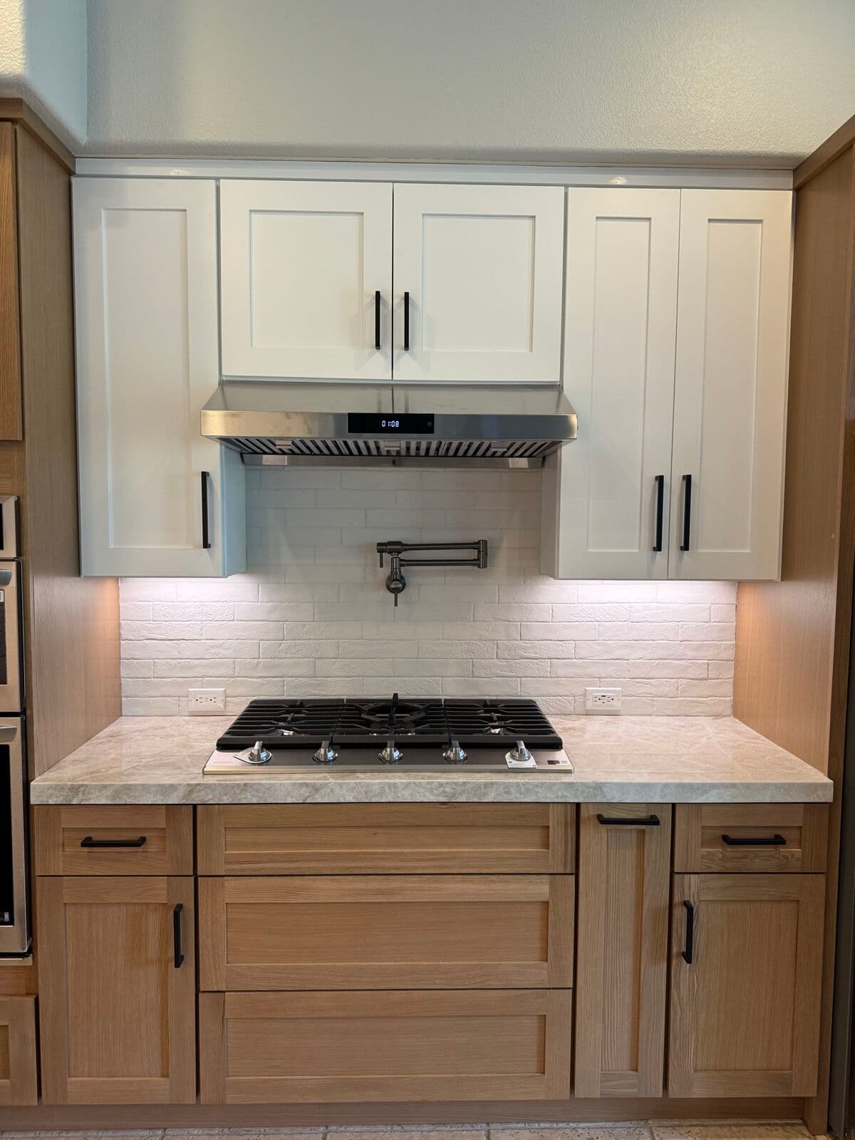 Gas cooktop wall with stainless under-cabinet hood, pot filler, white brick-look tile backsplash, white uppers and oak base cabinets