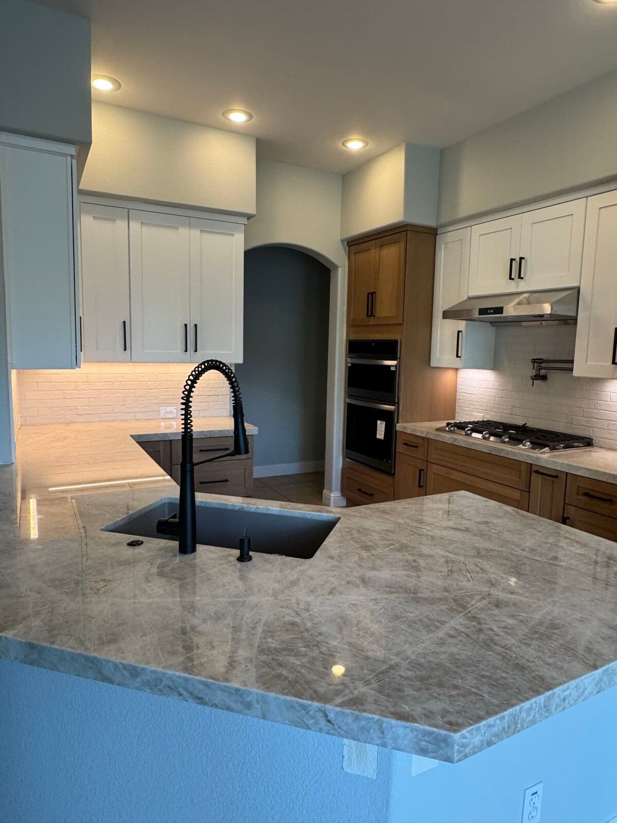 Close view of island stone countertop and black faucet with the oven cabinet, cooktop, and hood in the background