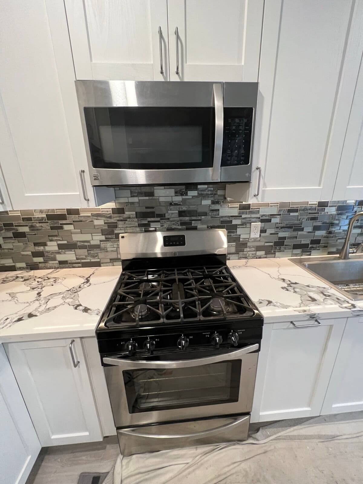 Stainless gas range and over-range microwave between white cabinets with gray mosaic tile backsplash