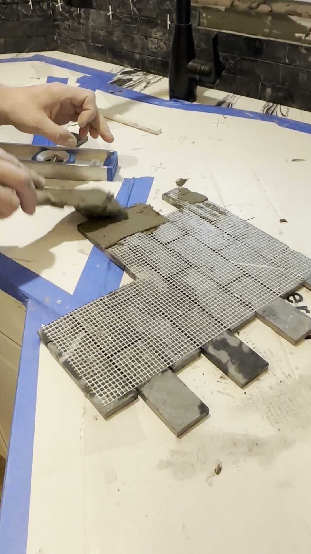 Hands back-buttering a mesh-backed black mosaic sheet on a protected kitchen countertop below a black faucet