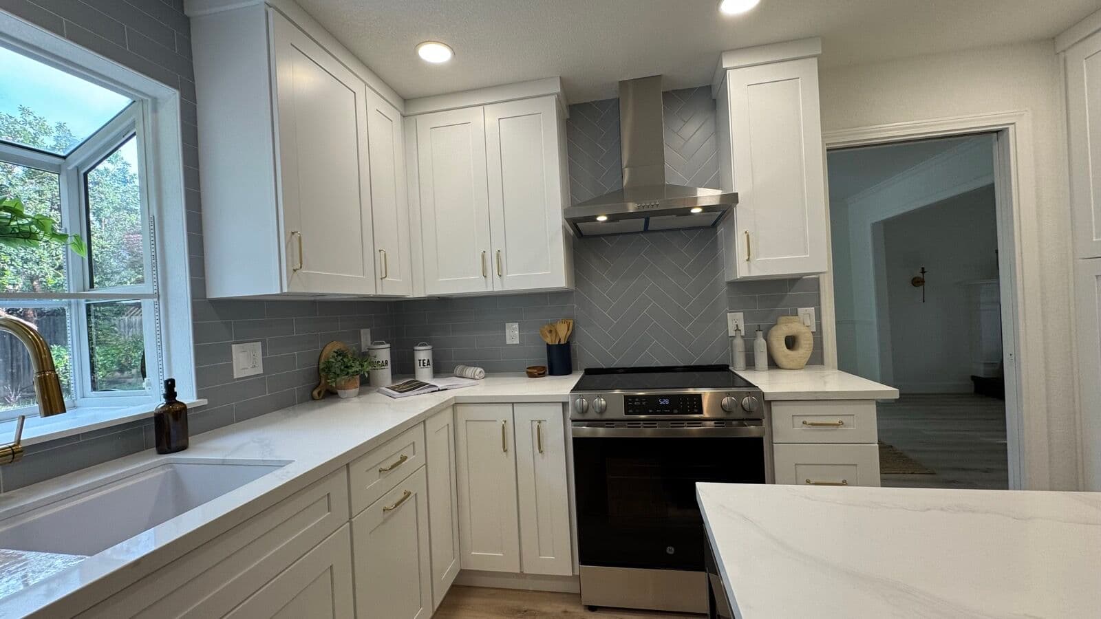 Kitchen sink wall with garden window, brass faucet, white cabinets, and stainless range under a chimney hood