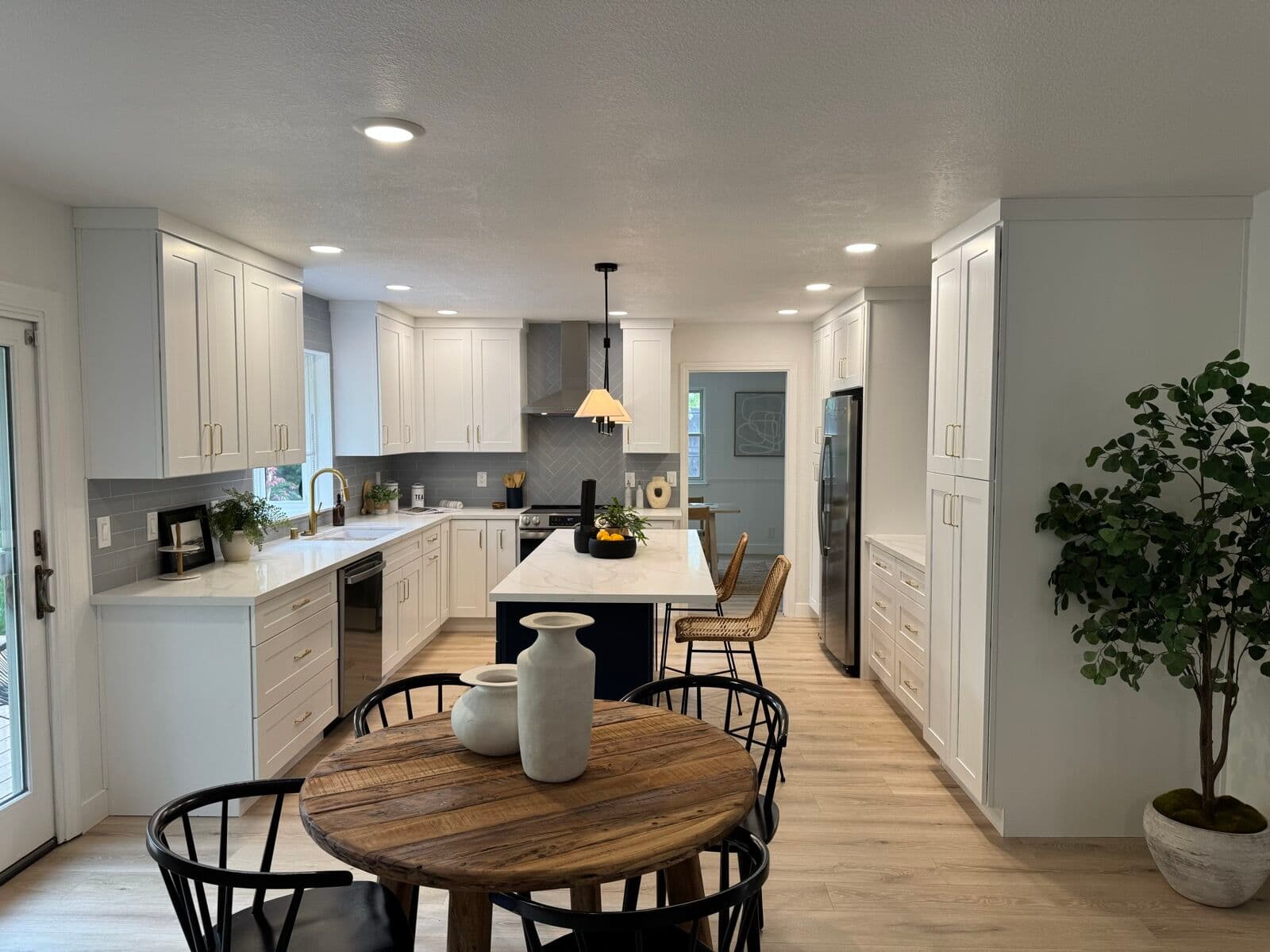 Wide view of white shaker kitchen with navy island, round wood dining table, and light plank flooring