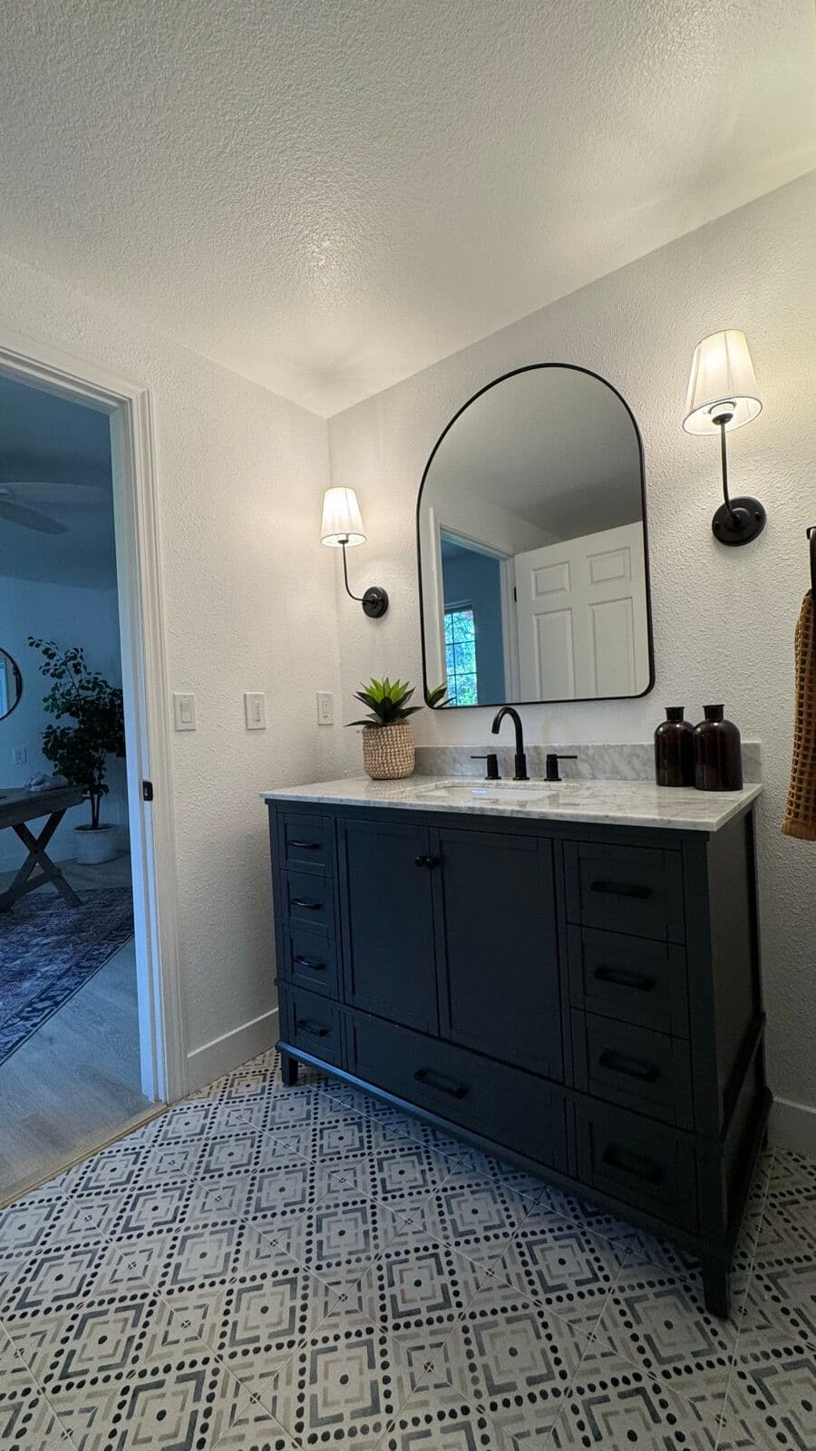 Hall bathroom with dark navy vanity, marble-look top, arched mirror, sconces, and patterned tile floor
