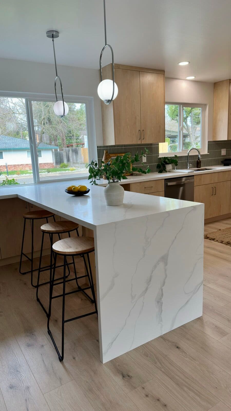 Waterfall quartz peninsula with wood bar stools in front of the kitchen sink wall and large windows