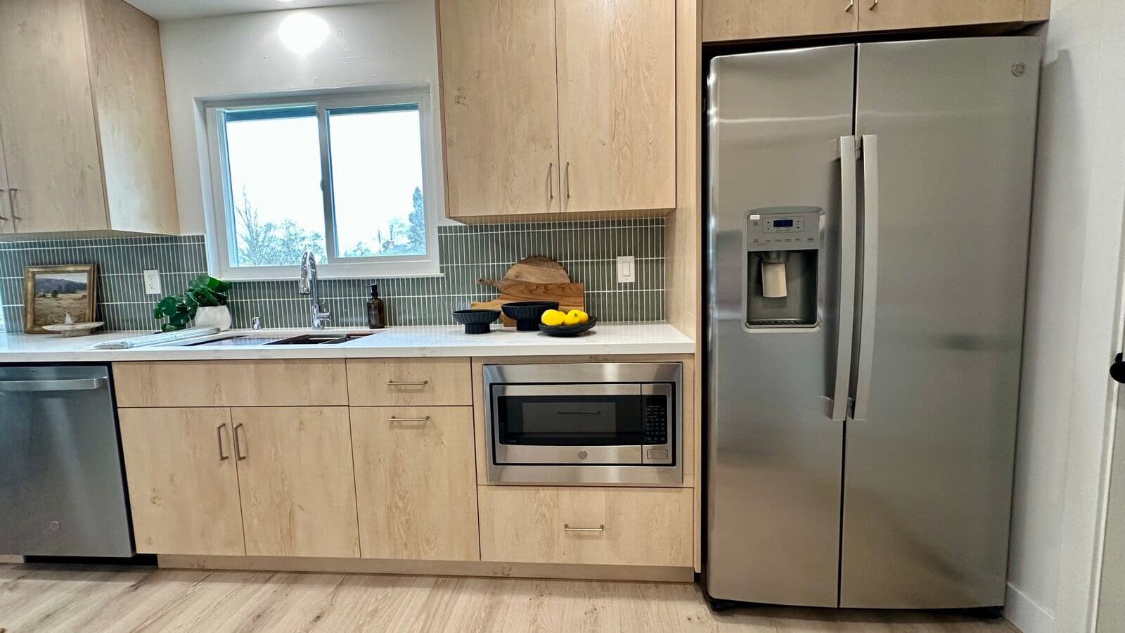 Kitchen sink run with window, sage stacked tile backsplash, built-in microwave, and stainless refrigerator
