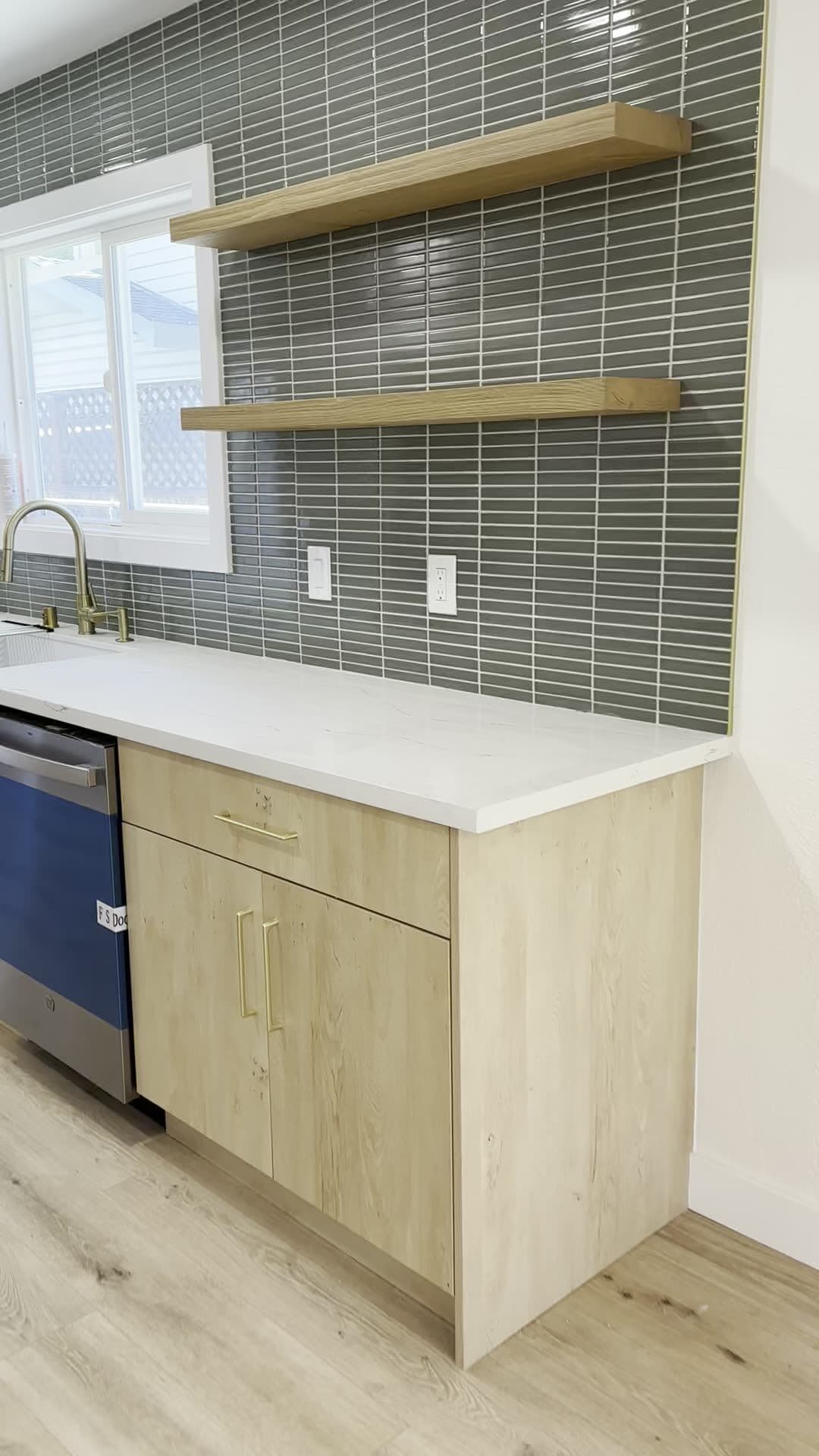 End of the cabinet run with a light wood base cabinet, brass pulls, white counter, and floating shelves on stacked tile above