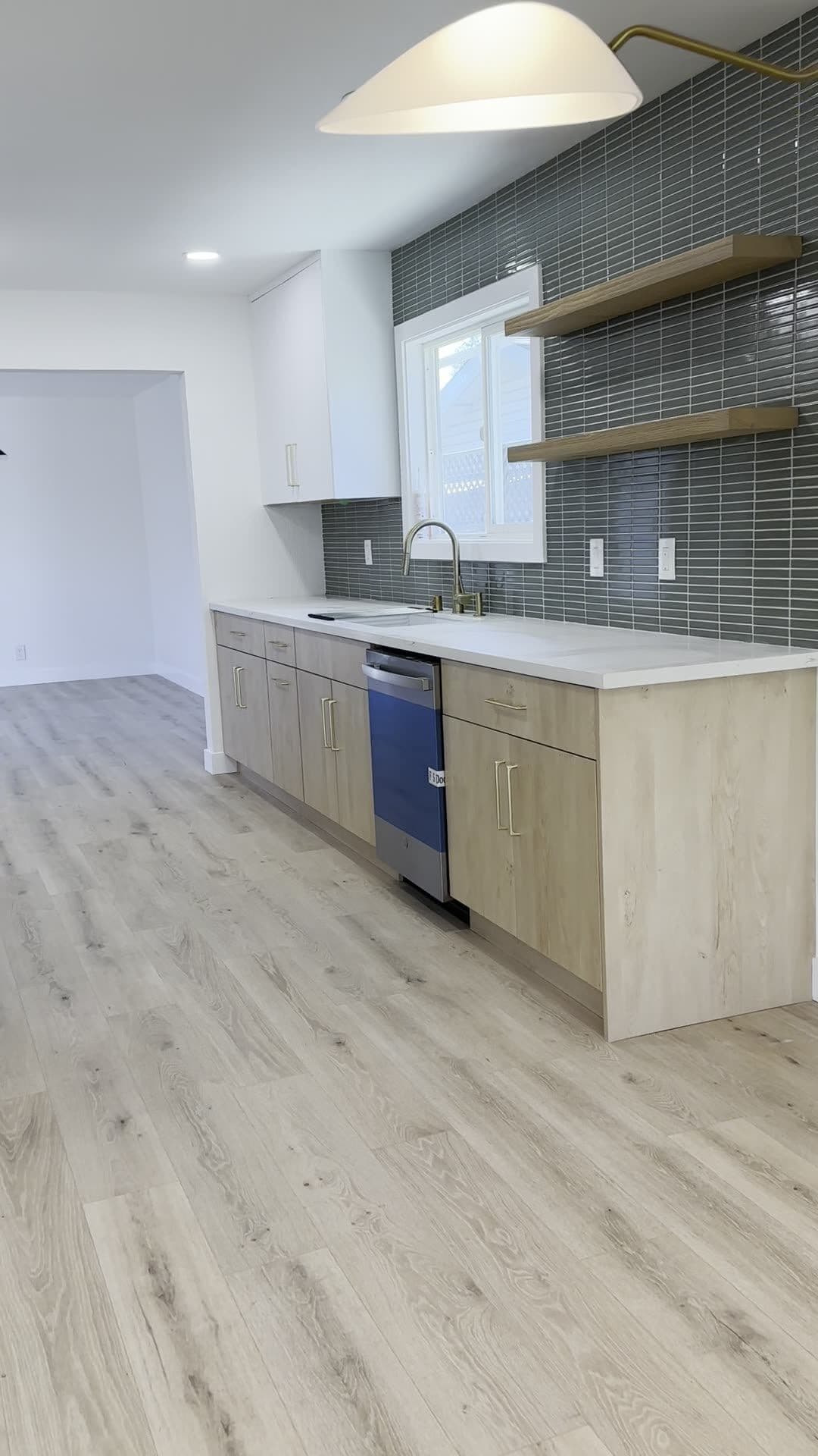 Wide view of the kitchen cabinet run with brass faucet, quartz-look counter, and wide-plank light flooring continuing into the next room