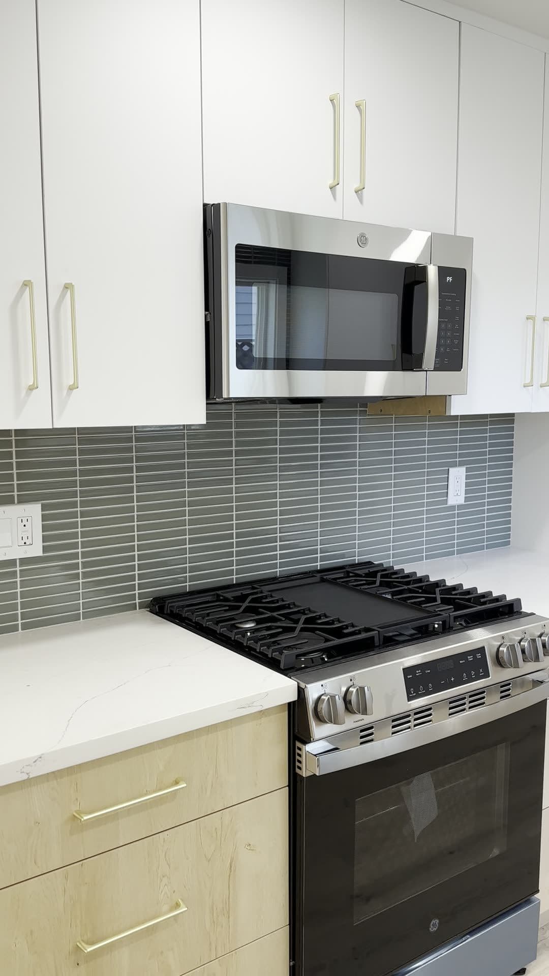 Stainless gas range and over-the-range microwave between white upper cabinets with brass pulls and a gray stacked tile backsplash