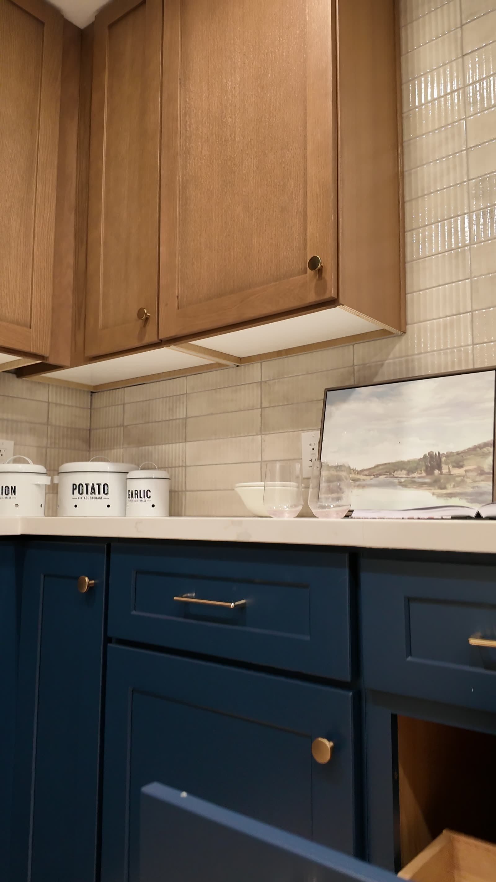Close view of a white-oak upper cabinet with brass knob over navy drawers and textured tile backsplash