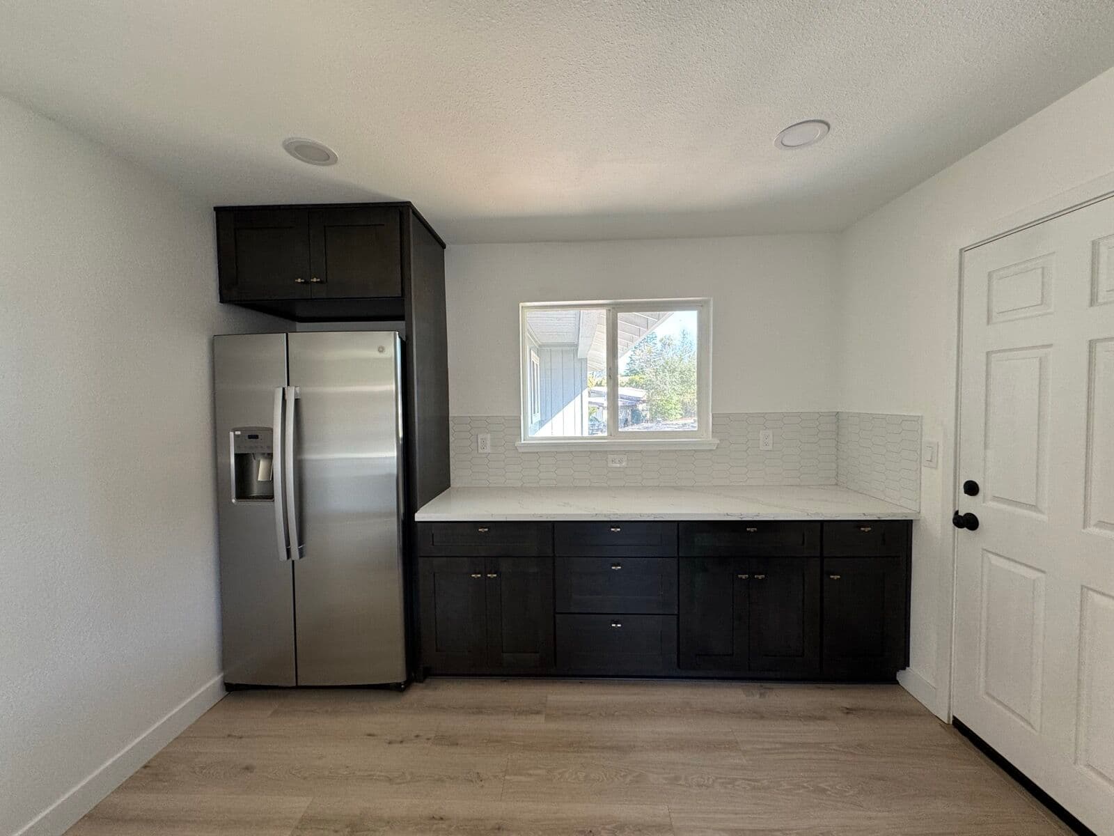 Dark base cabinets with white quartz counter and tile backsplash next to the stainless refrigerator and back door