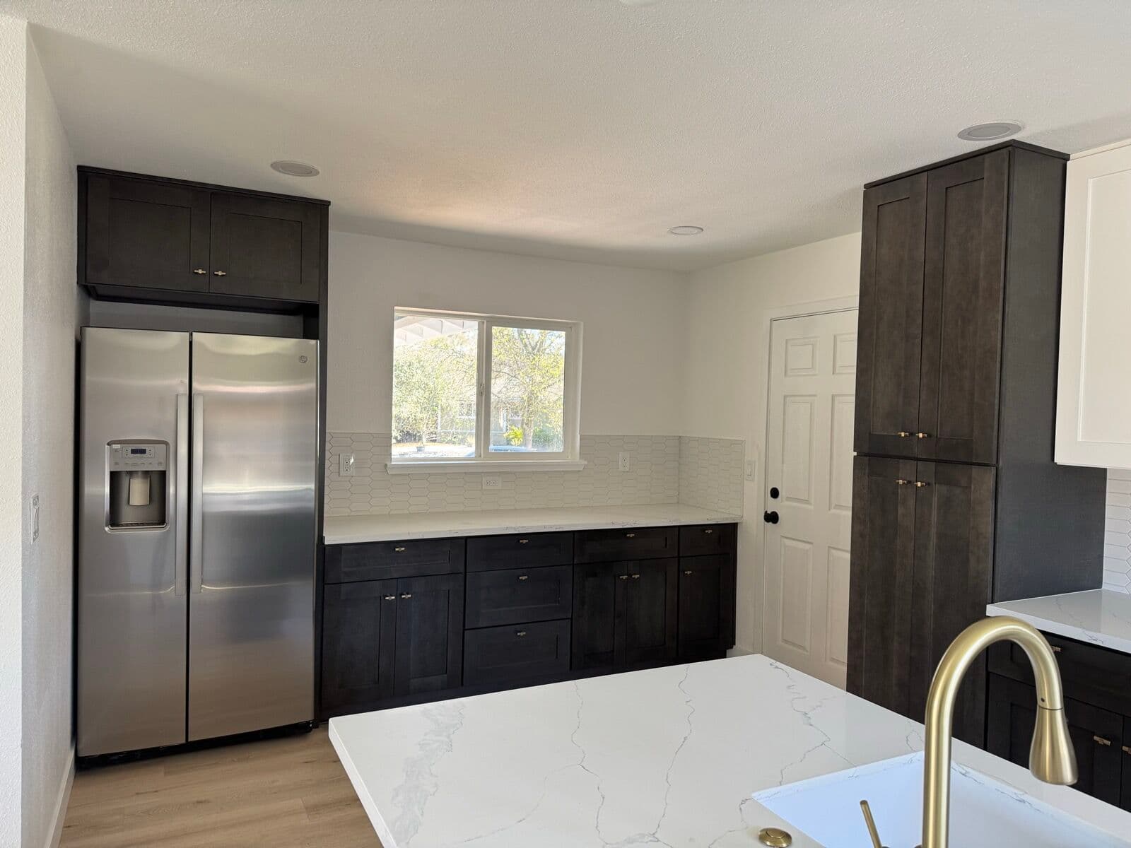 Stainless side-by-side refrigerator boxed in with dark cabinets beside a window and counter run