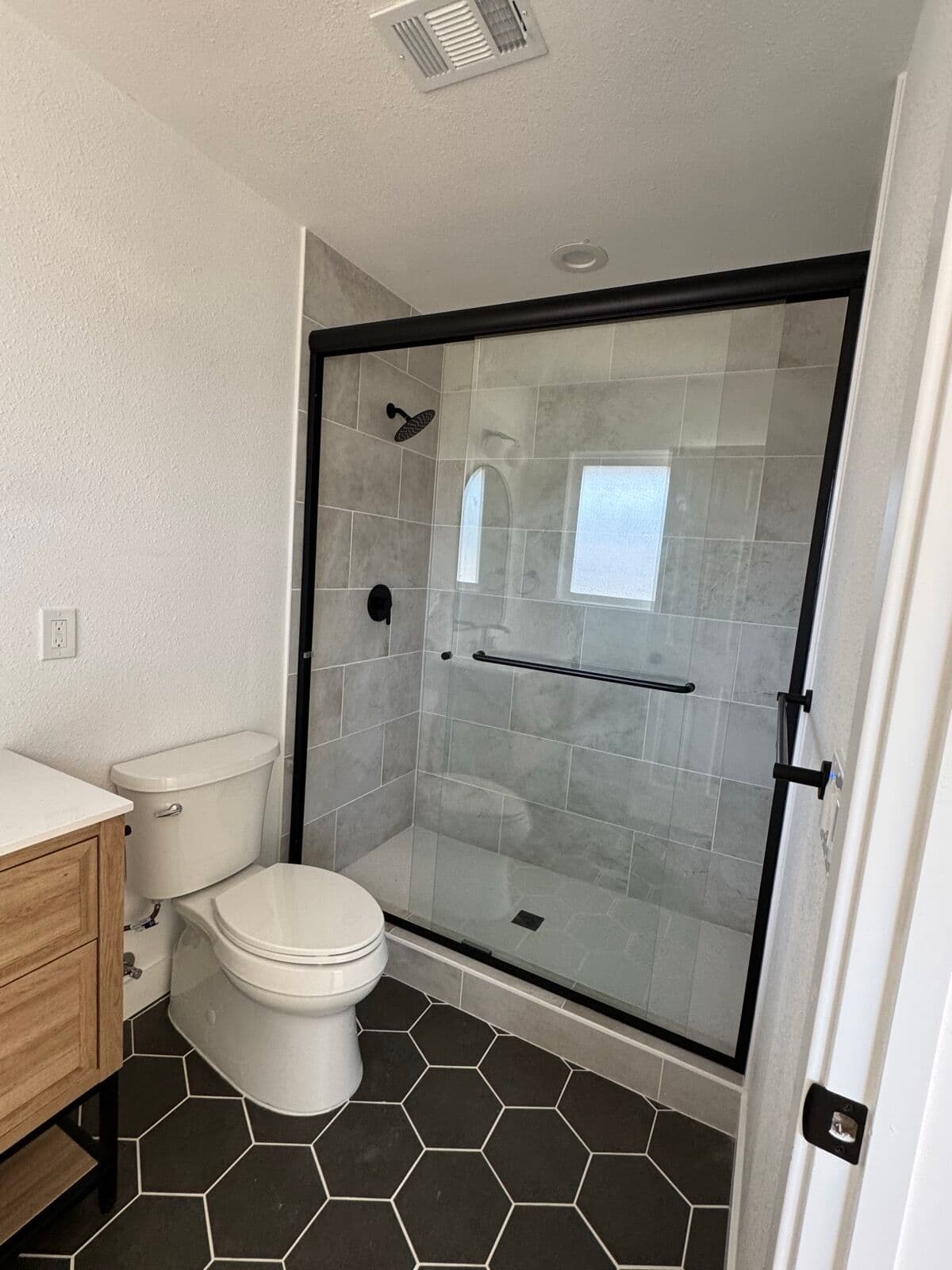 Walk-in shower with large-format gray tile, black rain shower head, and black-framed sliding glass door