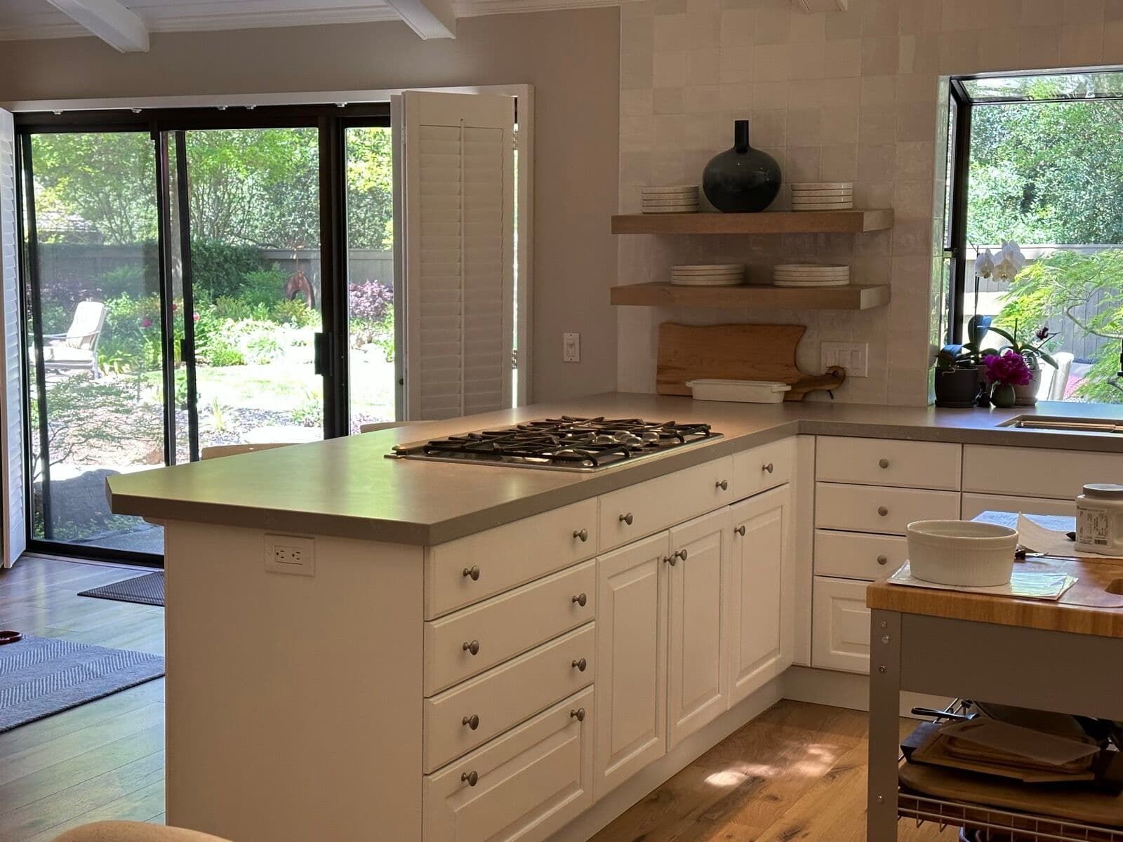 Cooktop peninsula with gray quartz counter, white raised-panel drawers, floating oak corner shelves, and sliding doors to the garden