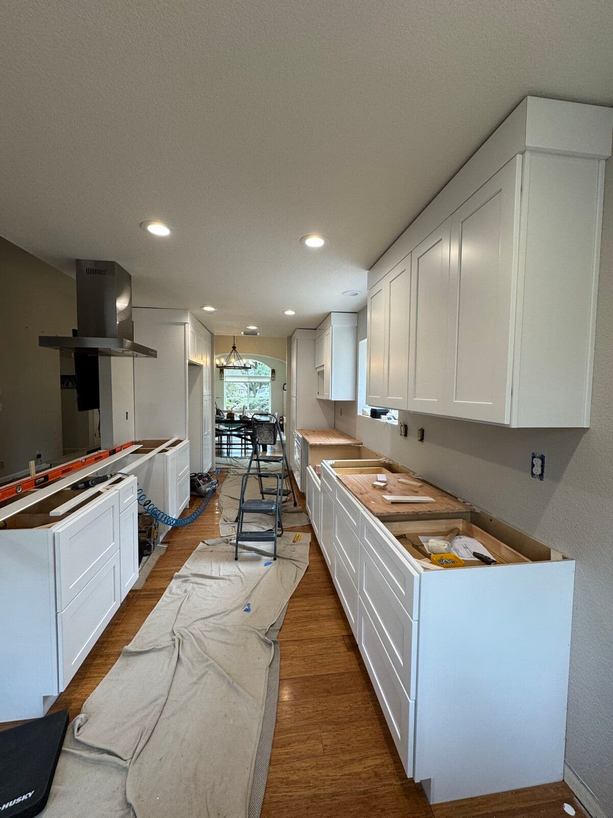 Wide view of a galley kitchen during cabinet installation with upper cabinets hung and base cabinets set on drop cloths