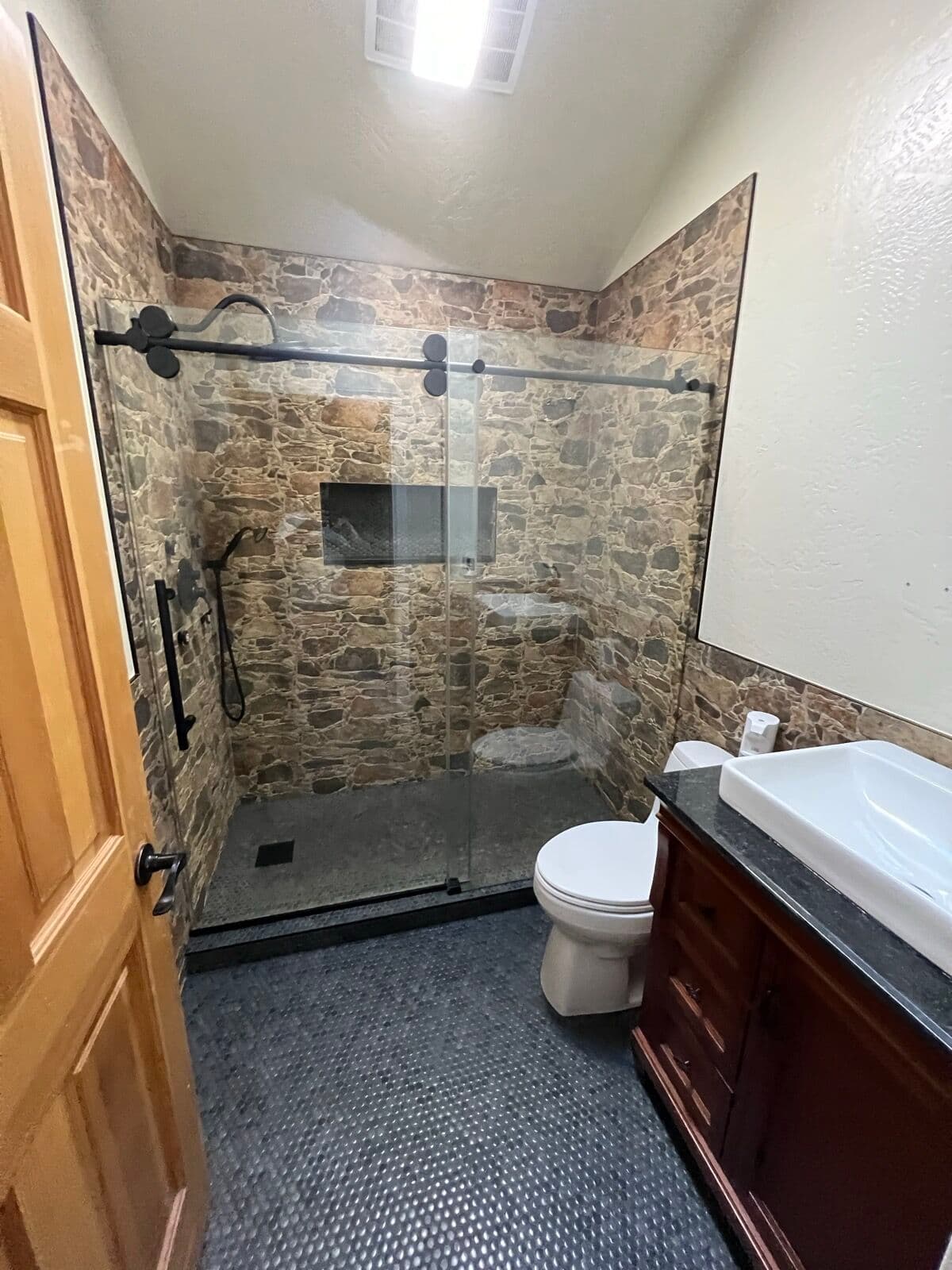 Bathroom with stone-look tile shower, black-frame sliding glass door, dark penny-tile floor, and dark wood vanity