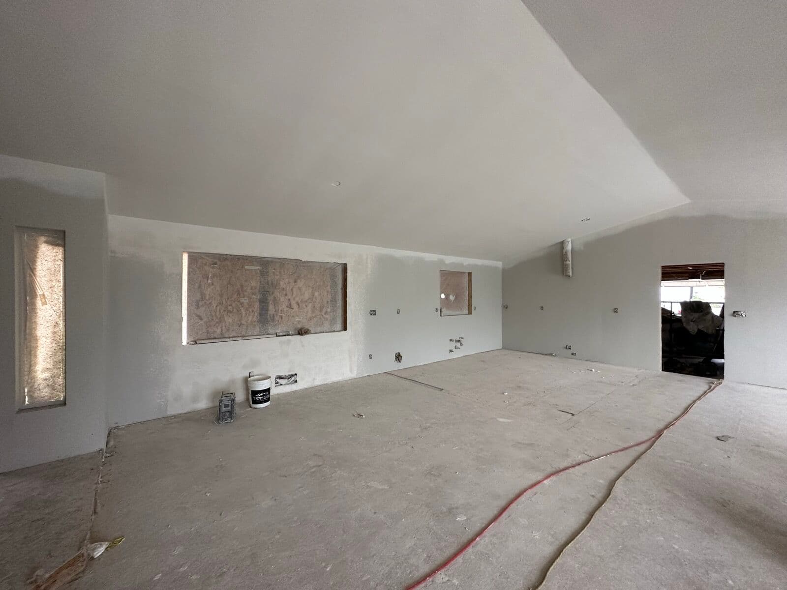 Before — Kitchen wall: primed drywall shell with boarded-up window openings and bare slab mid-build, finished out with white shaker cabinets, quartz counters, and a square tile backsplash.