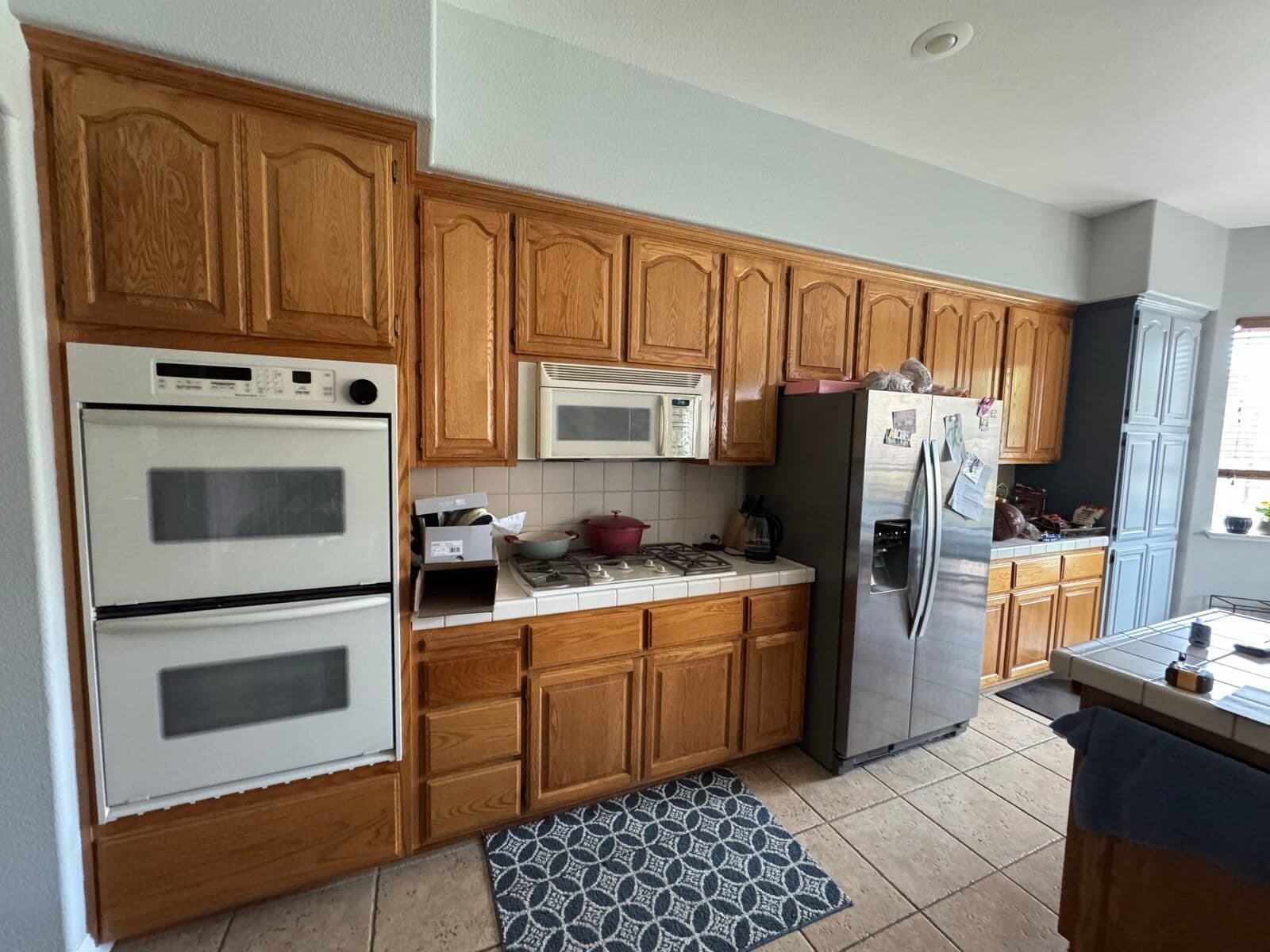 Before — Appliance wall: oak cabinets with white double oven and tile counters, rebuilt with an oak oven cabinet, gas cooktop with hood, and new refrigerator