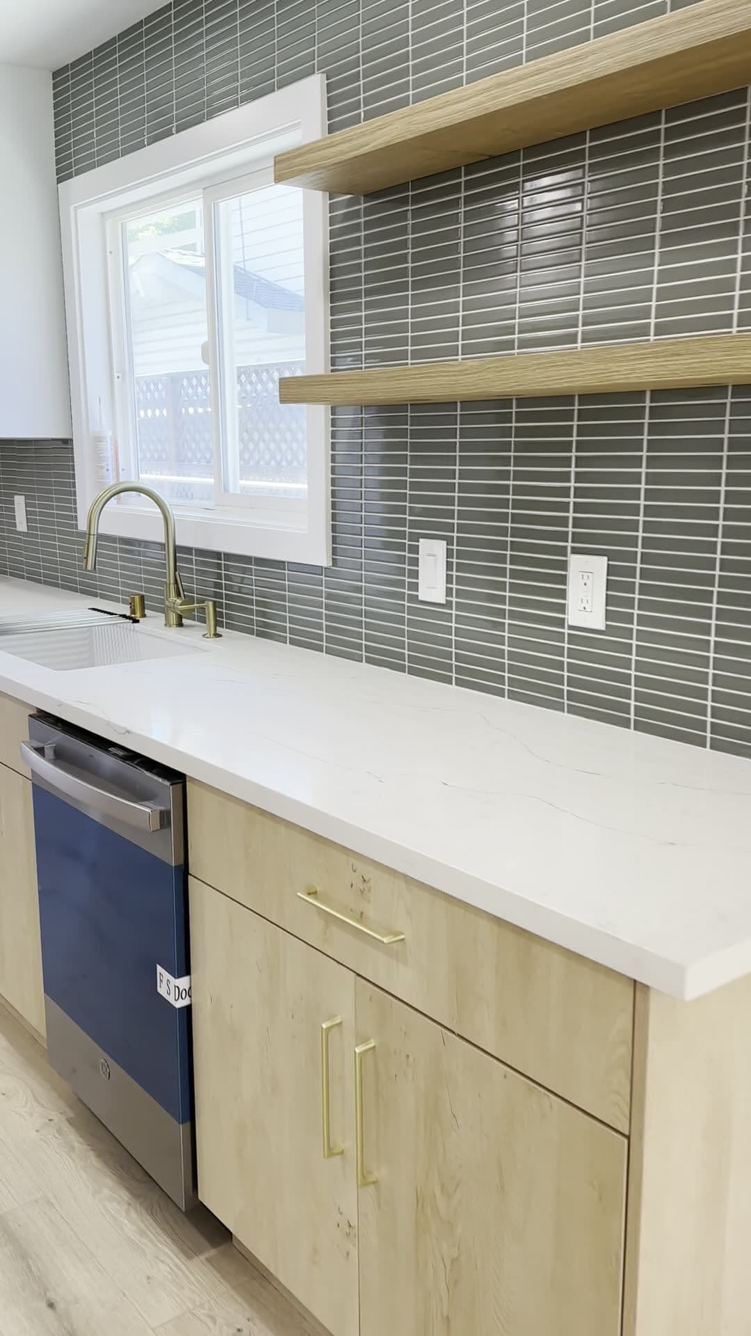 Two floating wood shelves mounted on a gray-green stacked tile wall beside a kitchen window