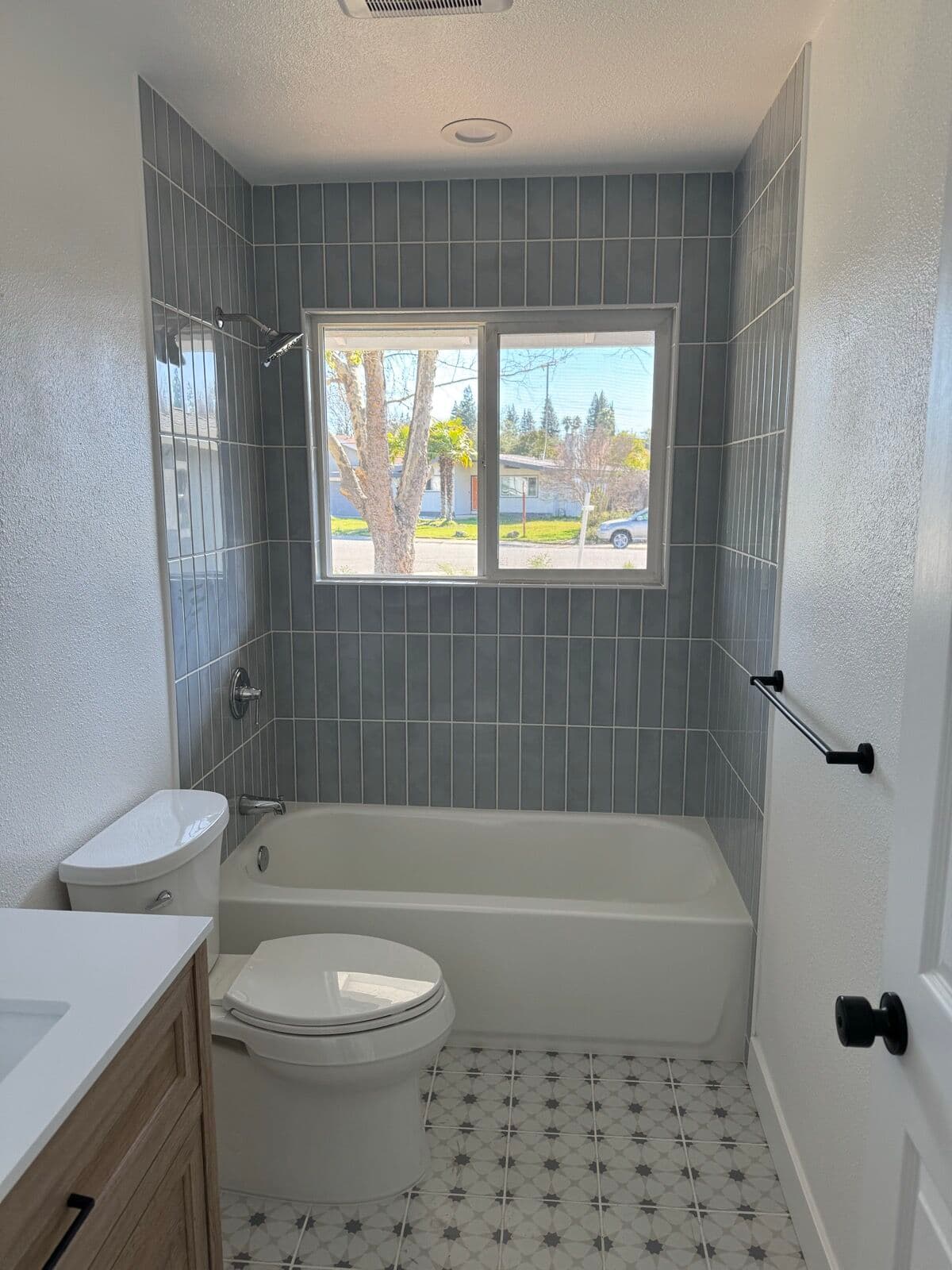 Tub with a gray-blue stacked-tile surround laid to the ceiling around a window