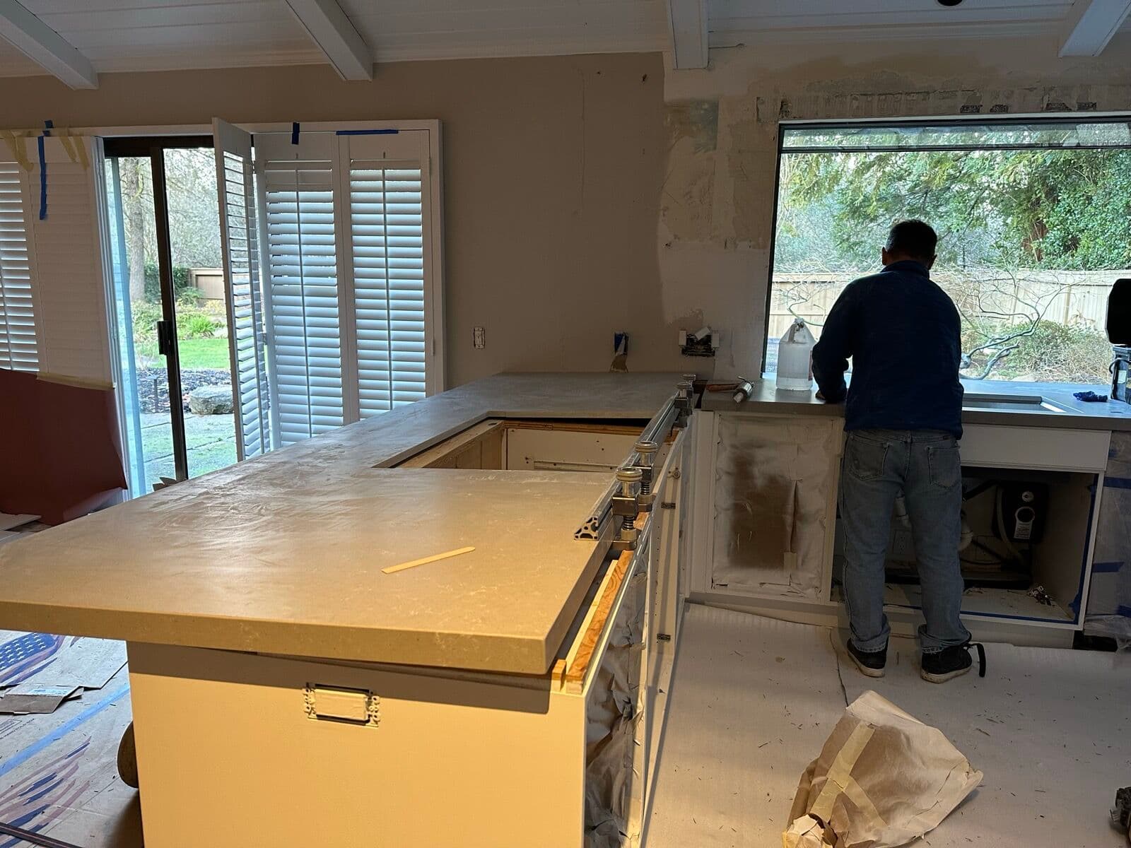 Before — Kitchen mid-remodel — walls stripped, cabinets masked off and the new quartz just set on the peninsula under the corner picture window — versus the finished kitchen with white cabinets, floating shelves and wide-plank oak floors.