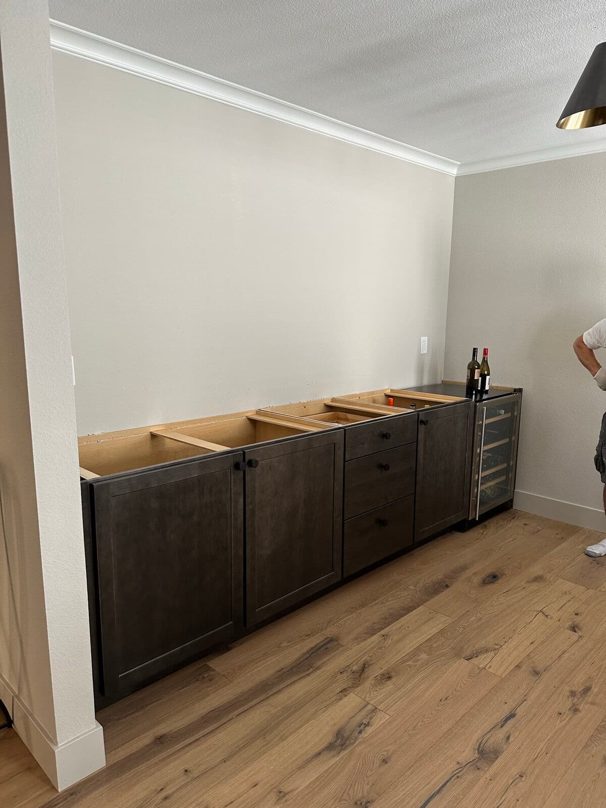 Before — Wine-bar wall mid-install — stained base cabinets and wine fridge set but still without tops — versus the finished run with its quartz counter and backsplash, shot from the same spot.