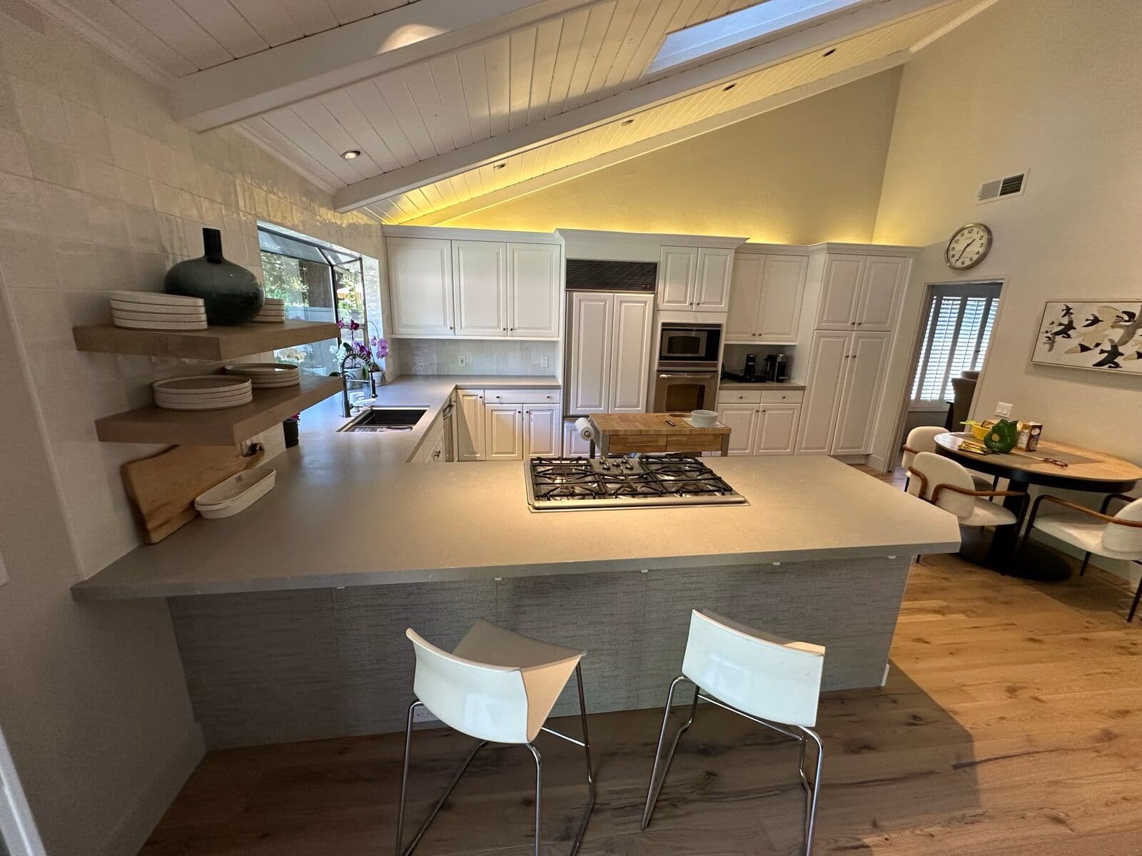After — Kitchen mid-remodel — walls stripped, cabinets masked off and the new quartz just set on the peninsula under the corner picture window — versus the finished kitchen with white cabinets, floating shelves and wide-plank oak floors.