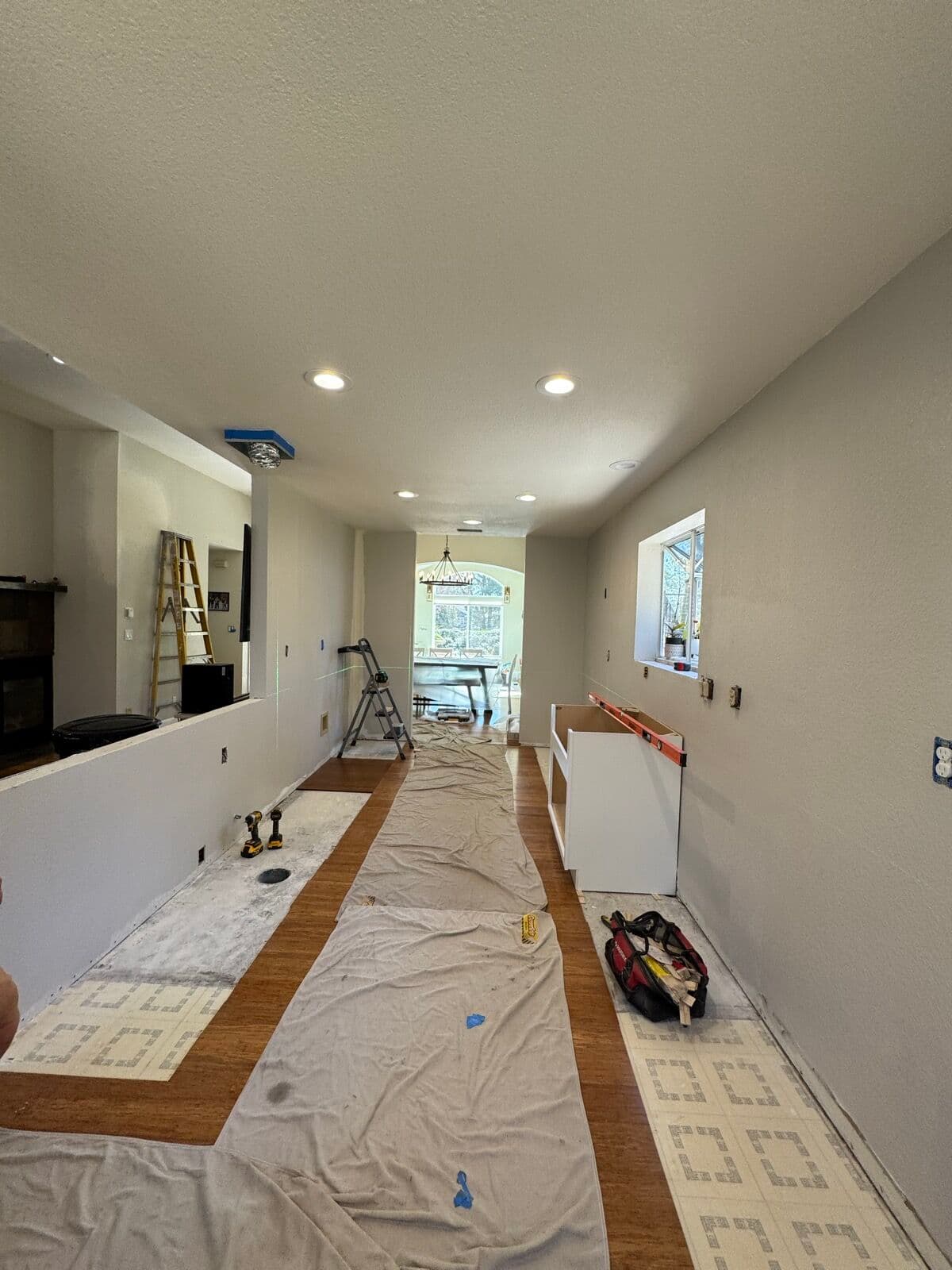 Before — Galley kitchen gutted to bare walls and old vinyl flooring with the pass-through half-wall still up and the first cabinet box staged, rebuilt with white shaker cabinets on both runs, a chimney hood, and an island where the half-wall came out.