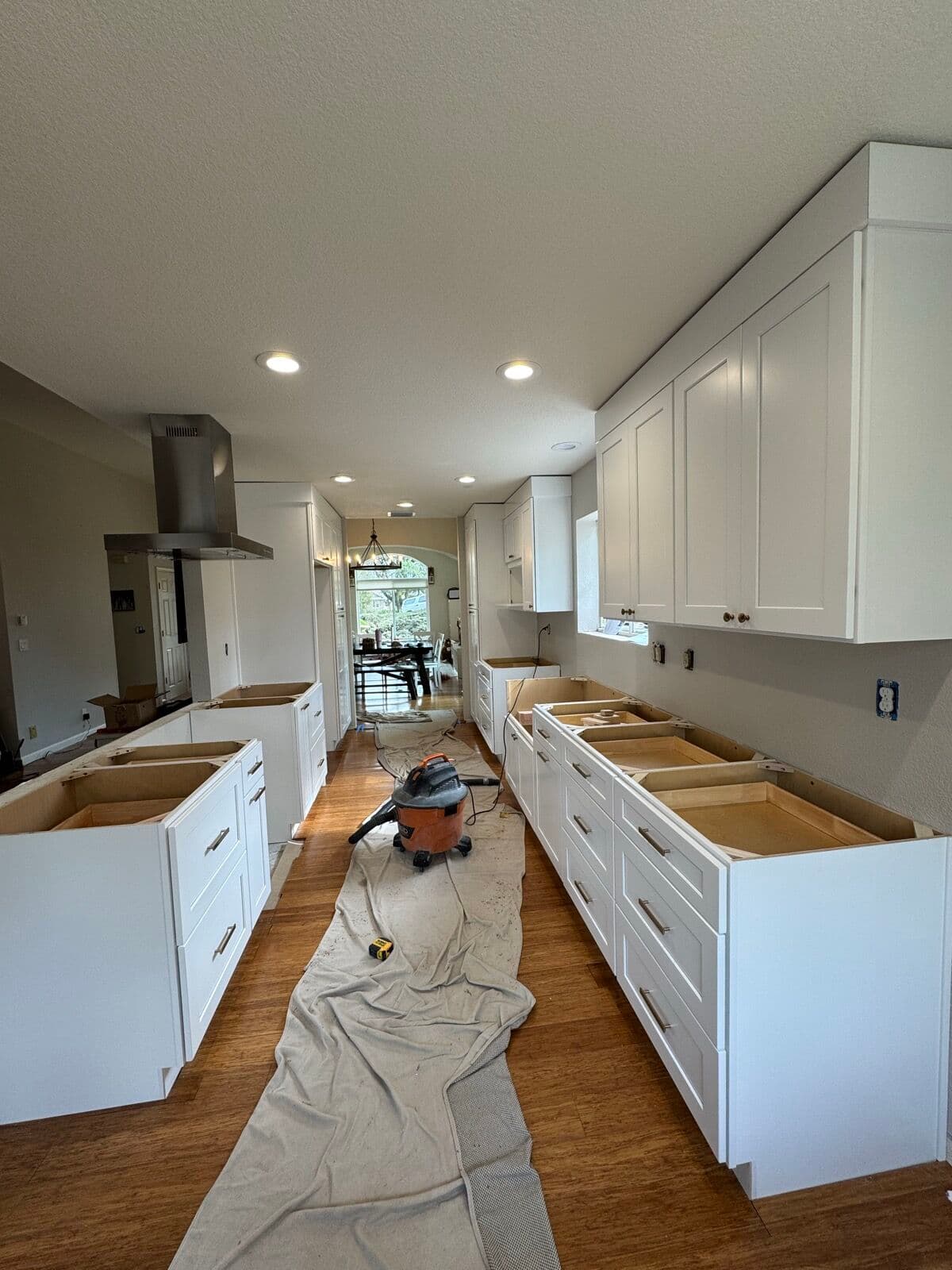 After — Galley kitchen gutted to bare walls and old vinyl flooring with the pass-through half-wall still up and the first cabinet box staged, rebuilt with white shaker cabinets on both runs, a chimney hood, and an island where the half-wall came out.